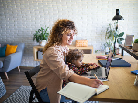Lächelnde Mutter mit Kleinkind auf dem Schoß. Am Schreibtisch mit Laptop arbeitend.