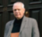 A white man with short white hair in a grey blazer is stood in front of a black door.