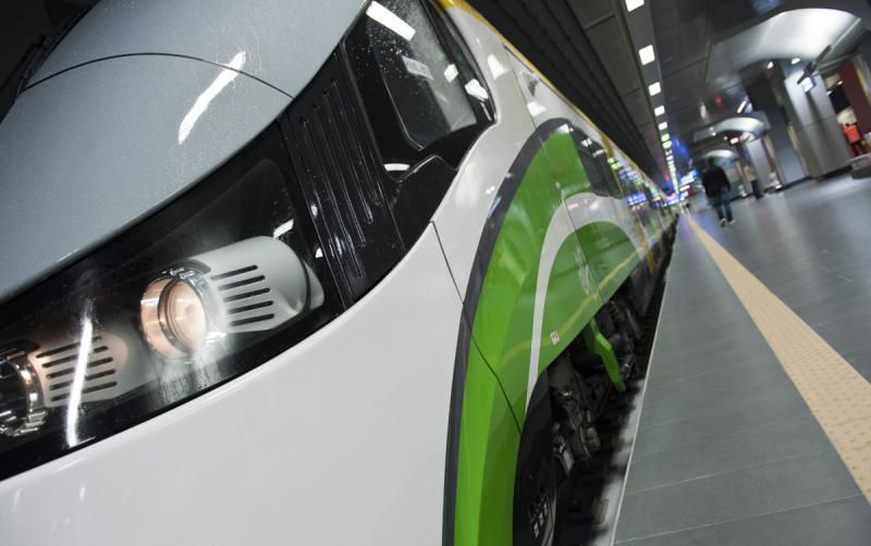 Przód nowoczesnego, białego pociągu z zielonymi pasami na boku, stojącego na stacji metra lub podziemnym przystanku kolejowym. W tle widać peron i sylwetki pasażerów.