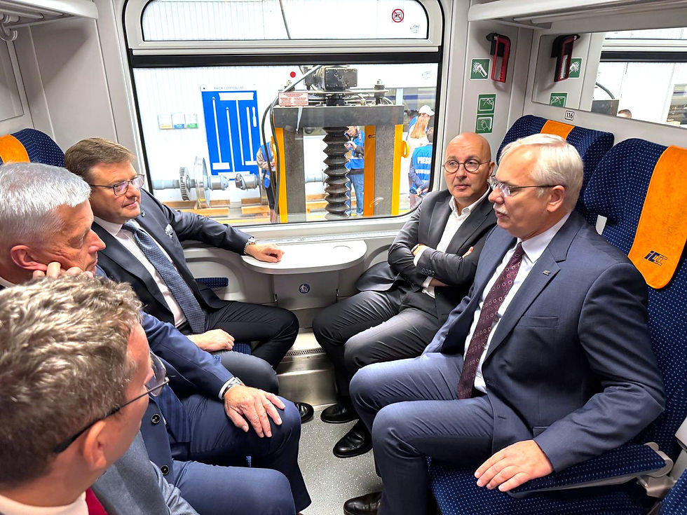 Czterech mężczyzn w garniturach siedzi w odnowionym przedziale wagonu kolejowego, a kolejny z mężczyzn znajduje się na pierwszym planie, obrócony tyłem do aparatu. Wszyscy rozmawiają, a w tle przez okno widać halę produkcyjną poznańskiej fabryki H. Cegielski - Fabryka Pojazdów Szynowych.