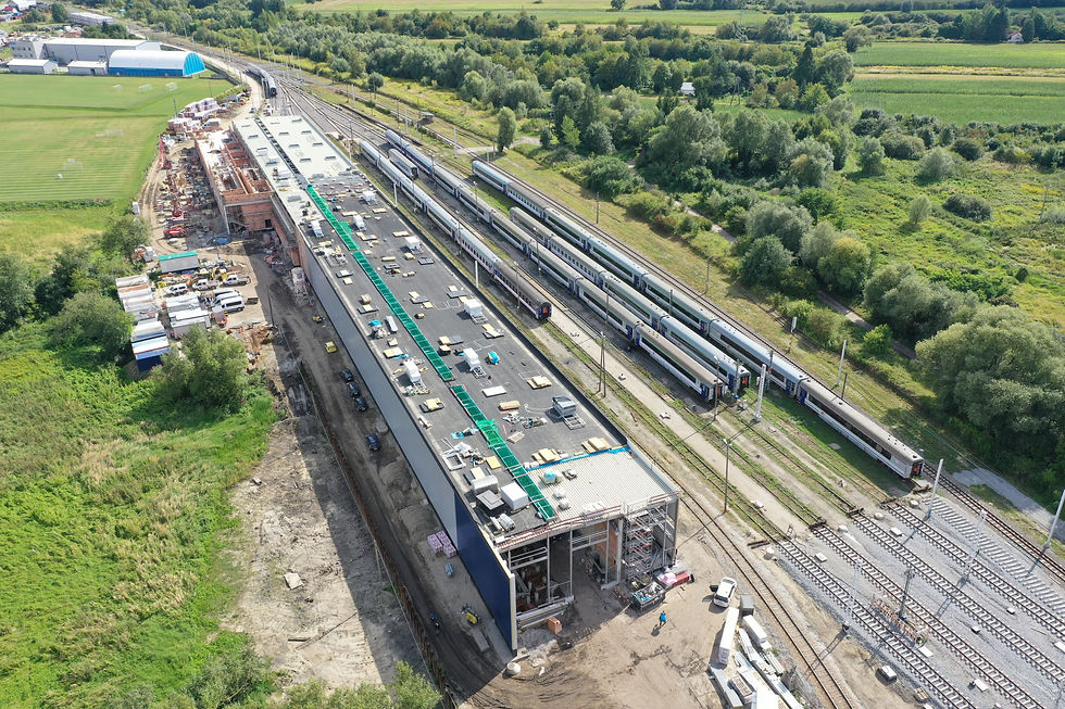 Na zdjęciu zrobionym z drona widać zaawansowane prace budowlane na terenie bazy technicznej PKP Intercity w Przemyślu. W centrum kadru dominuje długi, nowo wznoszony budynek hali napraw i myjni, z widocznym płaskim dachem oraz częściowo odsłoniętą konstrukcją. Obok budynku, po prawej stronie, na sąsiednich torach stoją pociągi, w tym składy typu ED74. Plac budowy otoczony jest zielonymi terenami i drzewami. Na pierwszym planie po lewej stronie widać zaparkowany sprzęt budowlany i materiały, a na dalszym planie kolejne, istniejące już obiekty kolejowe i rozległe tereny zielone.