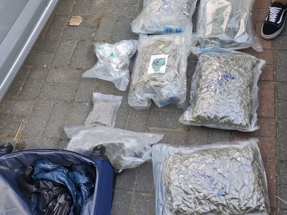 Zdjęcie przedstawia dużą ilość narkotyków, głównie suszu roślinnego, zapakowanych w przezroczyste, próżniowe worki foliowe. Worki są ułożone na szarym chodniku z kostki brukowej, obok niebieskiej walizki, z której część zawartości jest widoczna. W tle widać fragmenty pojazdu i stopy w czarno-białych butach sportowych.