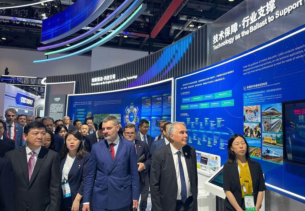 Prezes PKP S.A. Alan Beroud w granatowym garniturze idzie pośród innych uczestników, w tle widać nowoczesne stoisko wystawowe z grafikami i napisami.