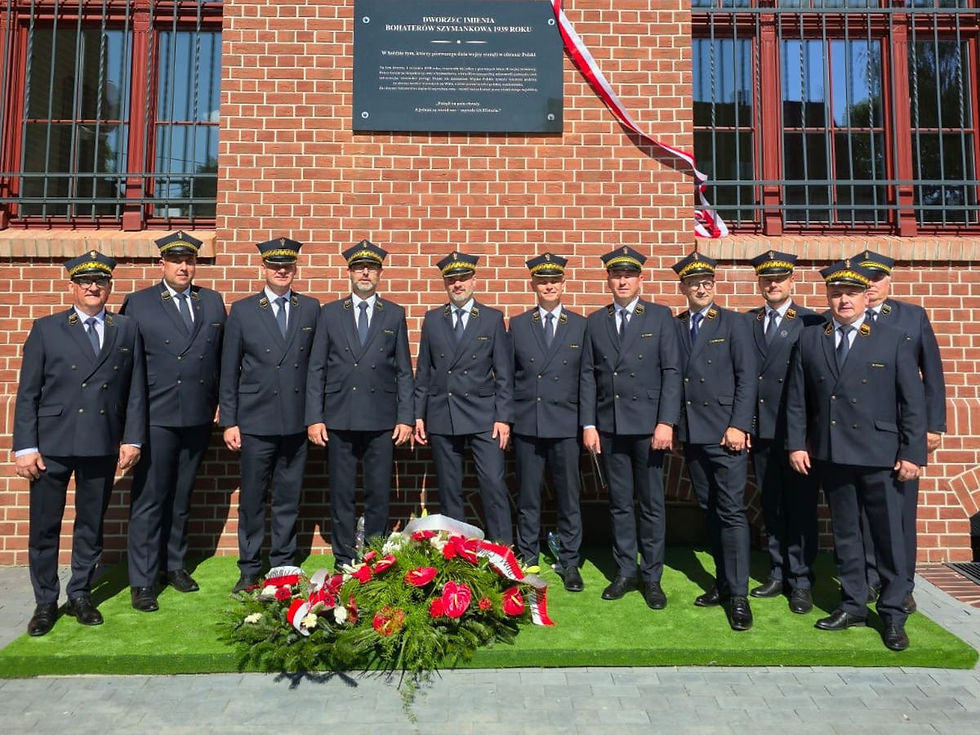Grupa umundurowanych kolejarzy stoi przed budynkiem dworca kolejowego, na którym znajduje się odsłonięta tablica pamiątkowa upamiętniająca Bohaterów Szymankowa z 1939 roku.
