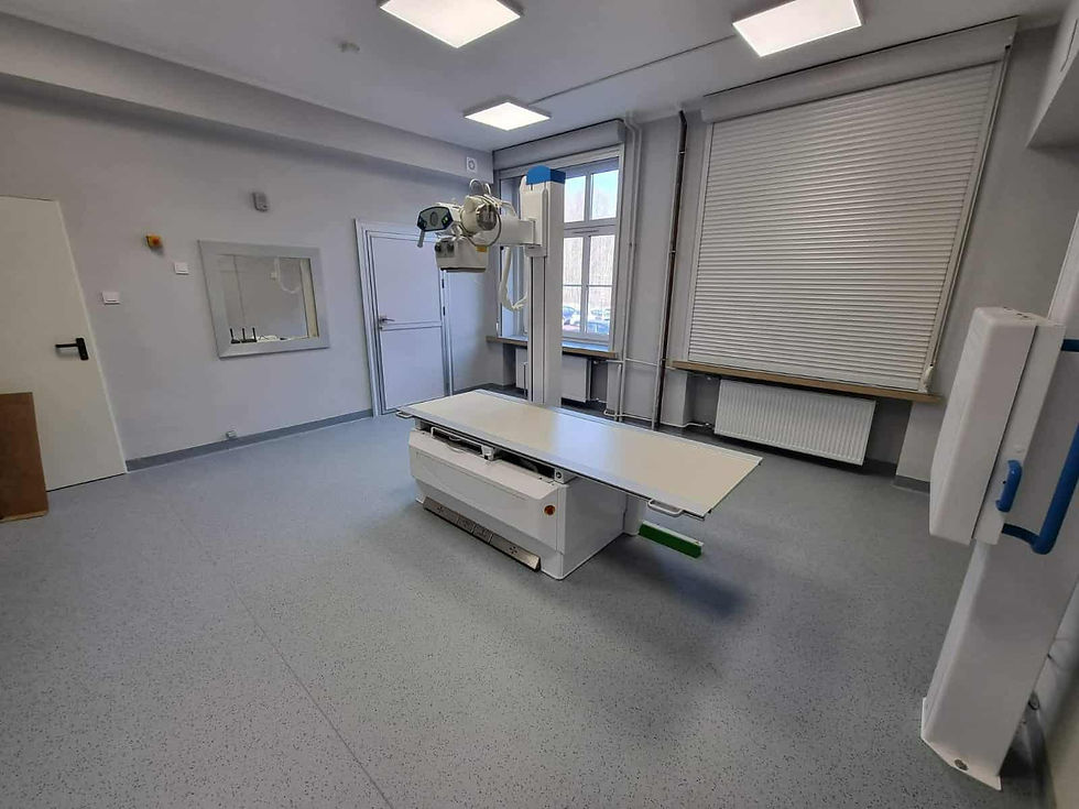 Nowoczesna pracownia rentgenowska z nowym, białym aparatem RTG umieszczonym centralnie na środku pomieszczenia, stołem do badań oraz fotelem biurowym w rogu, sugerująca czyste i świeżo wyremontowane wnętrze.