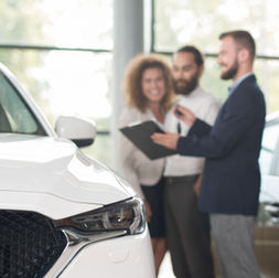 Consultor de concessionária conversa com casal ao lado de um carro branco, em ambiente de showroom, durante negociação de compra de veículo.