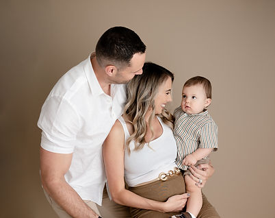 Mom and dad smiling at little boy while mom holds him and he looks at them. Family session in the mobile studio on pecan backdrop