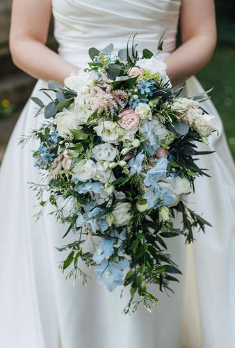 A cascading bridal bouquet featuring pastel blue delphiniums, white lisianthus, pale blush roses, jasmine and trailing greenery