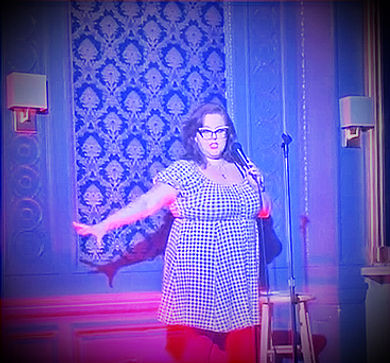 Audrey Liberman, a white woman in a vintage dress and cat-eye glasses, performing comedy.