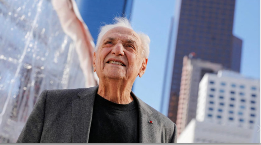 Frank Gehry in a gray blazer and black shirt looks up outdoors, with tall city buildings behind him.