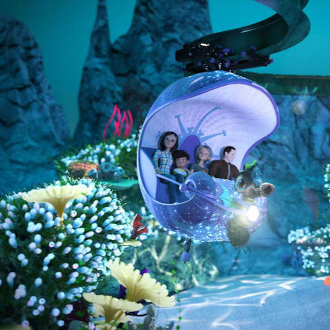 Four dolls ride a glowing purple shell pod through an underwater coral scene with blue rocks and soft light.