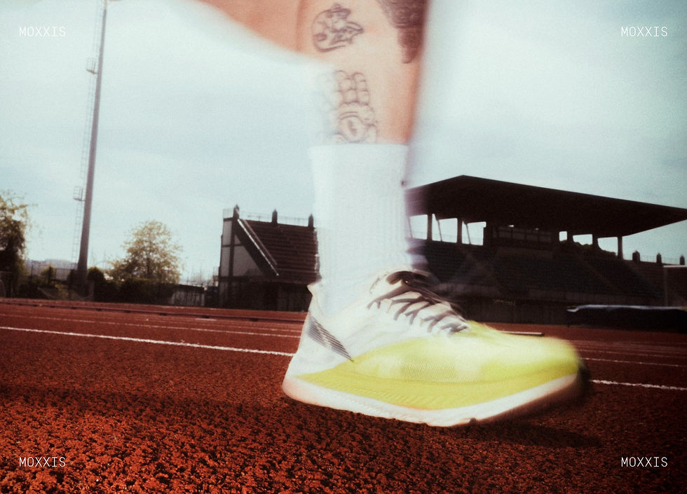 Bright yellow running shoe in motion on red track, blurred background of empty stadium seats. Visible tattoos on leg and "MOXXIS" text.