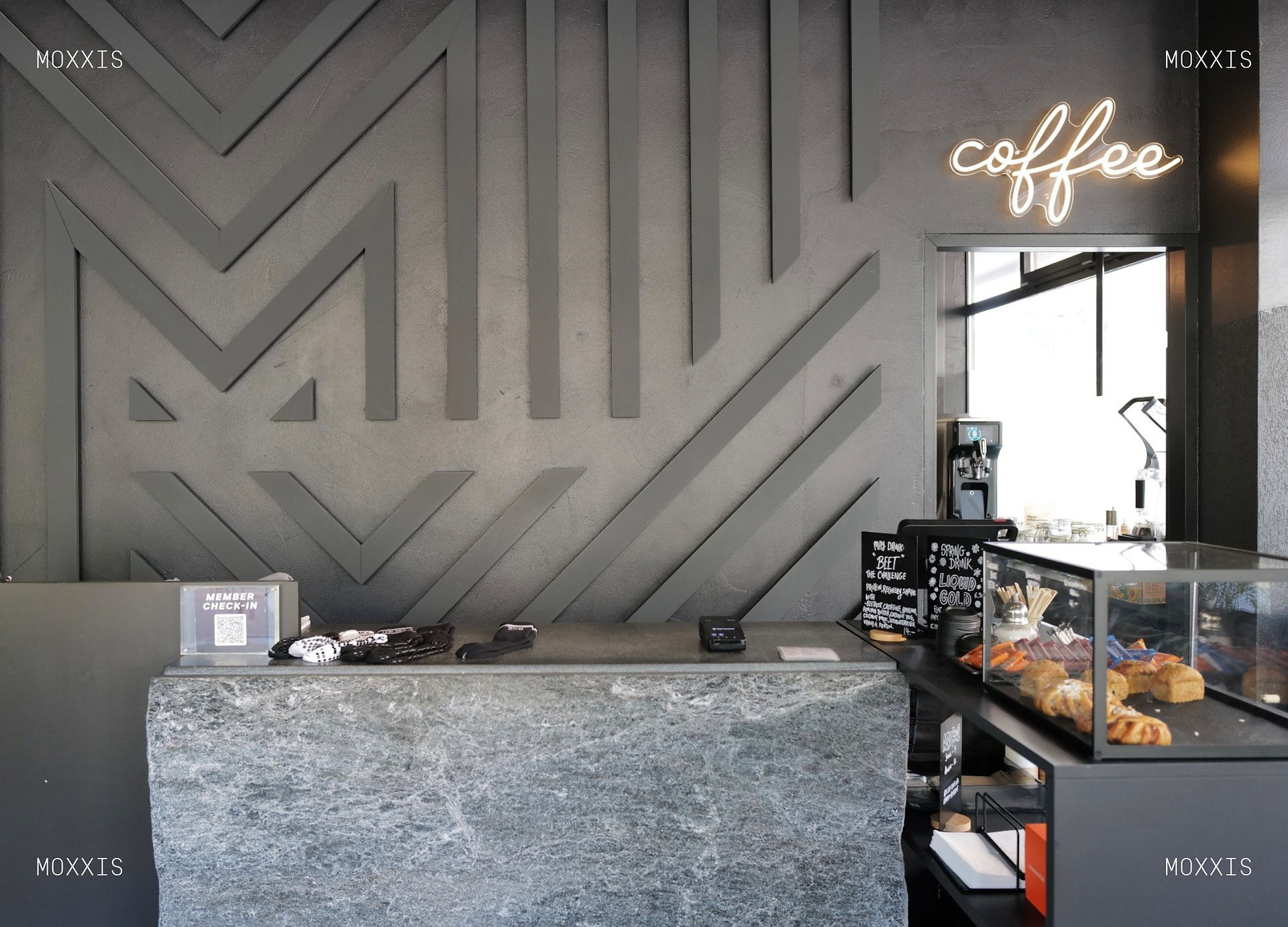  Reception area at The Mix sports studio in Seefeld. A raw natural stone counter with member check-in QR code, socks and gloves laid out for sale, and a small pastry case with croissants and snacks. Behind it, a dark gray wall with a geometric M pattern in relief and a glowing neon "coffee" sign. Chalkboards advertise the "Beet the Challenge" recovery shake and the "Liquid Gold" spring drink.