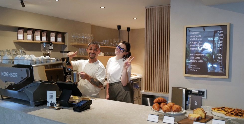 Two baristas behind the counter at Neighbor Coffee, one giving a thumbs up, the other a peace sign. Espresso machine, bags of coffee beans and a price menu in CHF in the background. Croissants and cakes lined up on the counter.