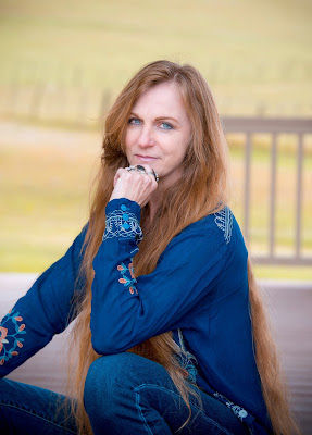 Author Sherri Dodd, a woman with long hair in blue shirt sits outdoors, resting her chin on her hand. Blurred background showing a fence. Calm and contemplative mood.