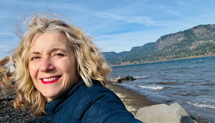 Hilary King smiles by a rocky coast in Oregon, wearing a dark jacket. Mountains and blue sky are visible in the background.