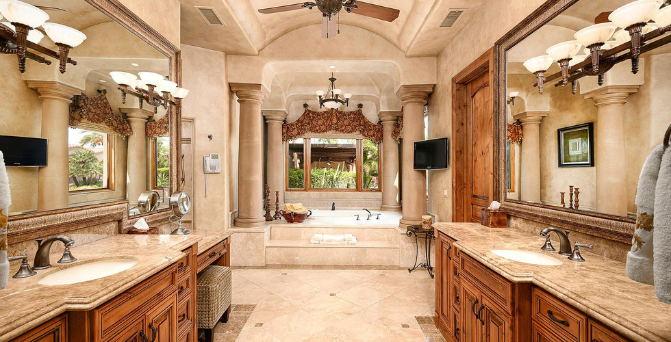 Luxury Mediterranean Warm Ambience Master Bathroom showcasing a front left and right side stained walnut wood cabinetry black hardware pulls vanities each with Taj Mahal Quartzite Stone Countertops have undermounted oval shape white sinks with medium oil stain nickel metal counter faucets. The left vanity end is a lower make-up vanity with a make-up vanity chair. On each vanity wall you can find a wide big copper texture framed mirror each with 2 moroccan style mounted 3 bulbs sconces. All walls and ceiling are light cream while the floor is matt crema marfil stone tiles. To the very centered back is an arched semi rectangle shape lower ceiling entrance with a two steps up to a ground built-in jacuzzi surrounded by 4 moroccan style 15 inches diameter capital tuscan columns. The very back wall has a 3 face window perspecting a green terrace garden. Above the jacuzzi you can find a moroccan style dark stained bronze metal 6 bulbs ceiling chandelier. To the front between vanities the ceiling has a ceiling brown fan. Right before entering the jacuzzi the right wall has a stained walnut wood door matches the vanities stained walnut wood cabinets and to the left side of that same door there is a wall mounted 36 inches Wide x 24 inches Height TV. Right before entering the jacuzzi; the left wall has a wall mounted white house telephone.
