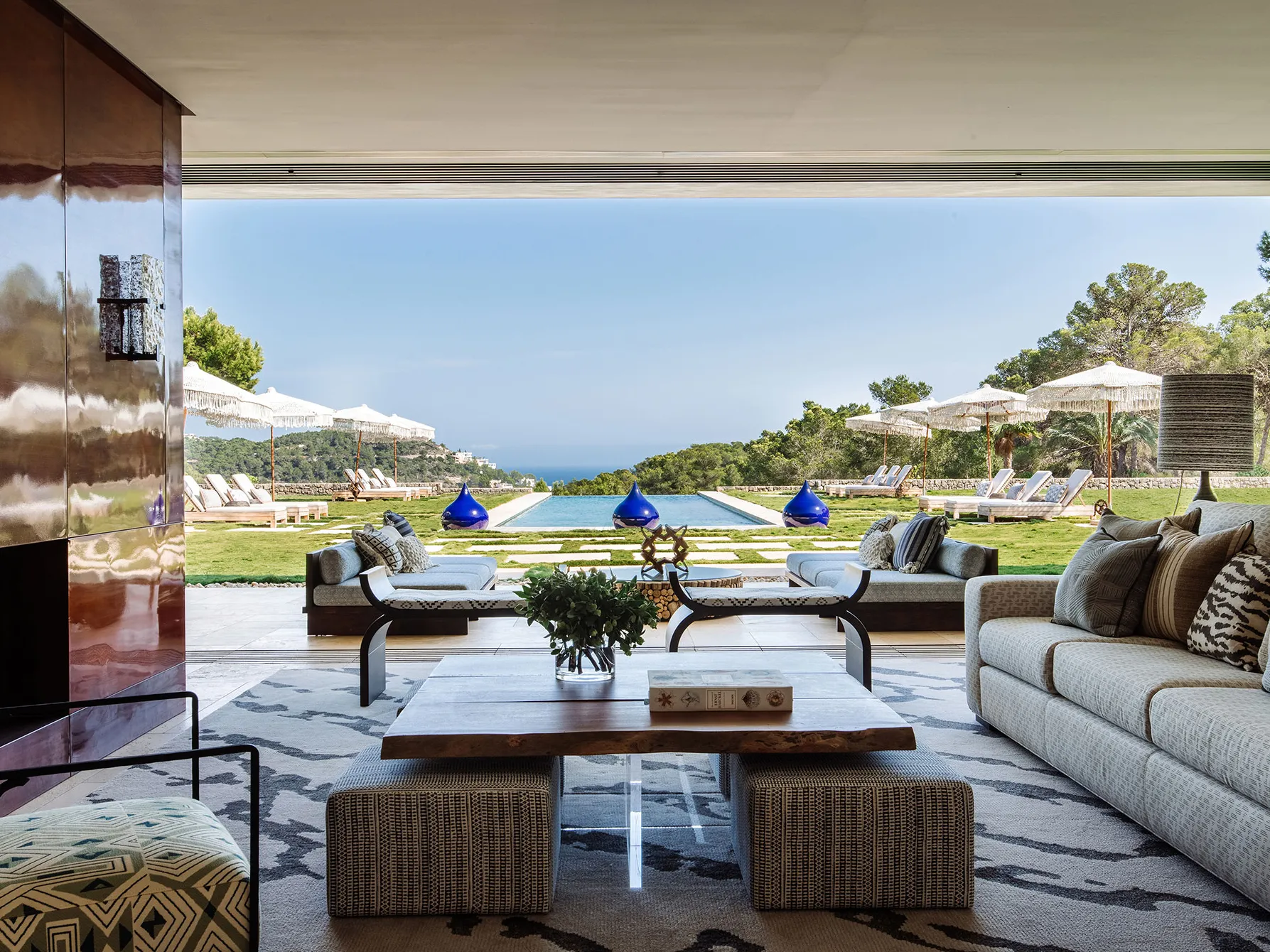 Luxury Beautiful Ibiza Architecture Interior Design Style Villa showing one of the many Open Concept Seating Furnitured Lounge Area openly connected to the Outdoor Nature Terrace has a green grass limestone tiling floor decorated with Ibiza Cream Cabanas Tents Beds and a Ground Built-In Infinity Pool. The Outdoor Terrace surroundings are filled with Nature Green Trees and Plants with a Background Blue Panoramic Sky. At center of image where the Terrace and beginning of pool you can see 3 perfectly positioned big deep bright blue moroccan vases.