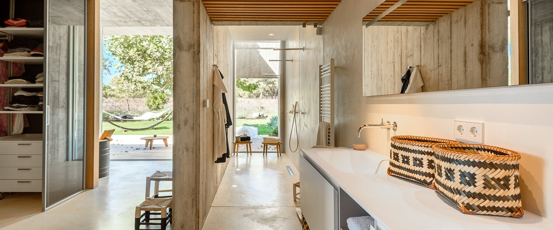 Luxury Ibiza Warm Ambience Master Bathroom with a near left side area has a Single Interior Partition Concrete Wall and Partial Ceiling, Interior Unfinished Limestone Floor and Remaining Walls. To the right hand side you can see a Cream Vanity with a Wall Hung Mirror has an Oiled Maple Wood Striped Ceiling Area. The far back is an Open Concept Terrace showing Green Grass Floor, Green Trees and Back Nature Brown Mountains. The are a couple of short wood chairs around.