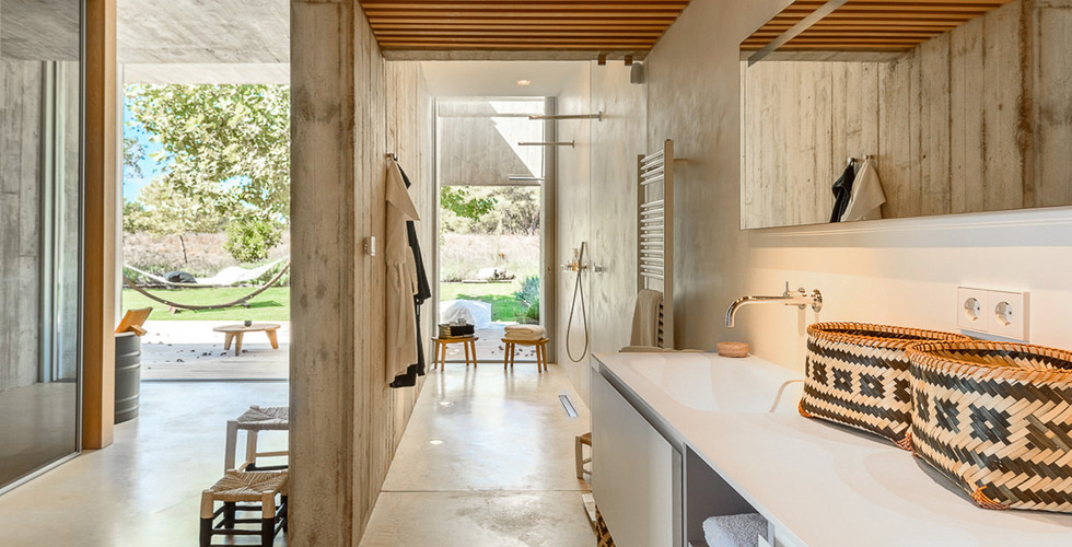 Luxury Ibiza Warm Ambience Master Bathroom with a near left side area has a Single Interior Partition Concrete Wall and Partial Ceiling, Interior Unfinished Limestone Floor and Remaining Walls. To the right hand side you can see a Cream Vanity with a Wall Hung Mirror has an Oiled Maple Wood Striped Ceiling Area. The far back is an Open Concept Terrace showing Green Grass Floor, Green Trees and Back Nature Brown Mountains. The are a couple of short wood chairs around.