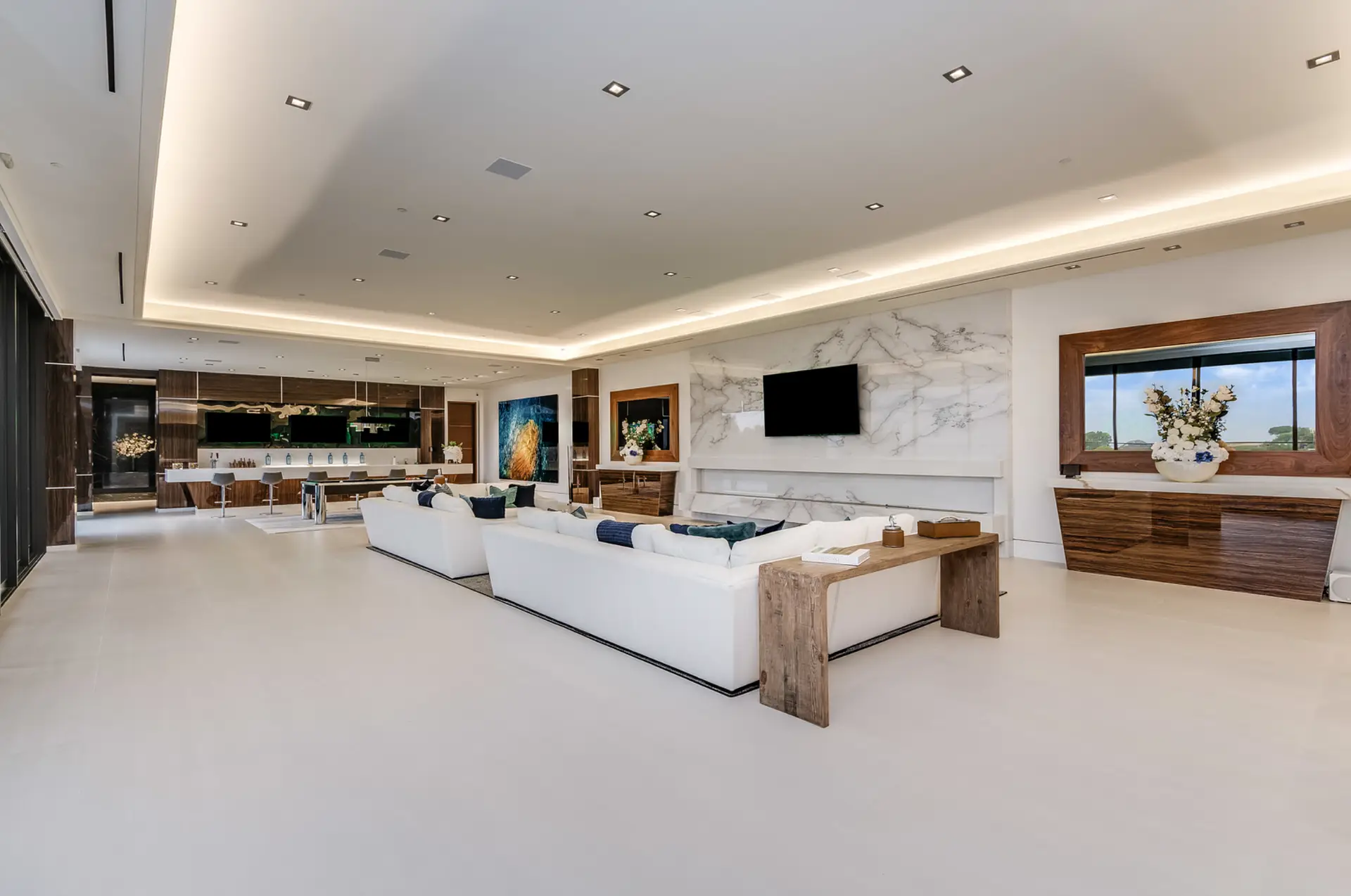 Ultra Modern High End Rectified Mega Mansion Interior Living Room area composed of 2 White Extra Large L-Shape Couches have a front 15 Feet Wide x 10 Feet Height x 4 Inches Thick High Gloss Calacatta Marble Stone has 1 Wall Mounted 6 Feet Wide x 5 Feet Height TV. To the right side and left side of the 15 Feet Wide x 10 Feet Height x 4 Inches Thick High Gloss Calacatta Marble Stone is a High Gloss Tiger Eye Geometrical Rectangled Console topped with a White Flowers Bouquet and a Wall-Mounted Big Framed Mirror. The back area has a High Gloss Tiger Eye Cabinetry Bar and an Additional 8 Seaters Seating Table. The entire floor is Matt Crema Marfil Tiles. The Ceiling is Multi-Leveled with 1 foot difference and has Ceiling Built-In Recessed Cans Warm Lighting System. Back far right wall has a Huge Wall Mounted High Gloss Glass Abstract Art Paint.