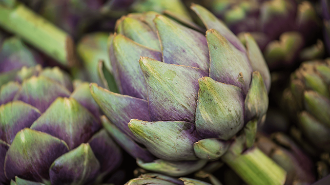 Close up of a green and purple artichoke on a pile of additional artichokes of the same colors.