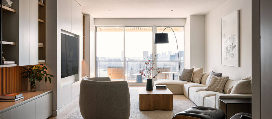 Modern living room with beige sofa, armchair, and black floor lamp. Large TV on the wall, city view through balcony windows, calm ambiance.