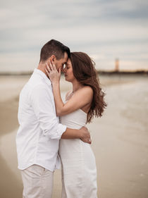 couple kissing standing on beach with lighthouse in background standing in sand