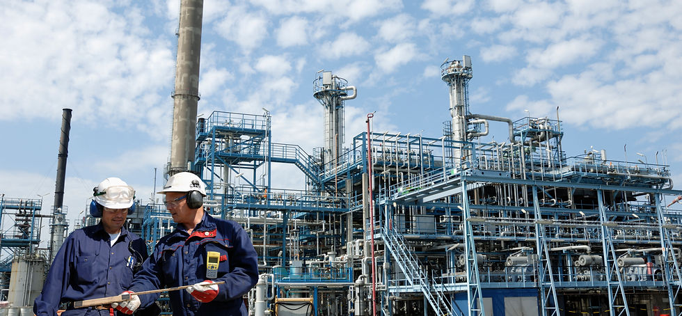 Two workers in blue uniforms and hard hats discuss a plan at an industrial plant, with metal structures and a blue sky in the background.