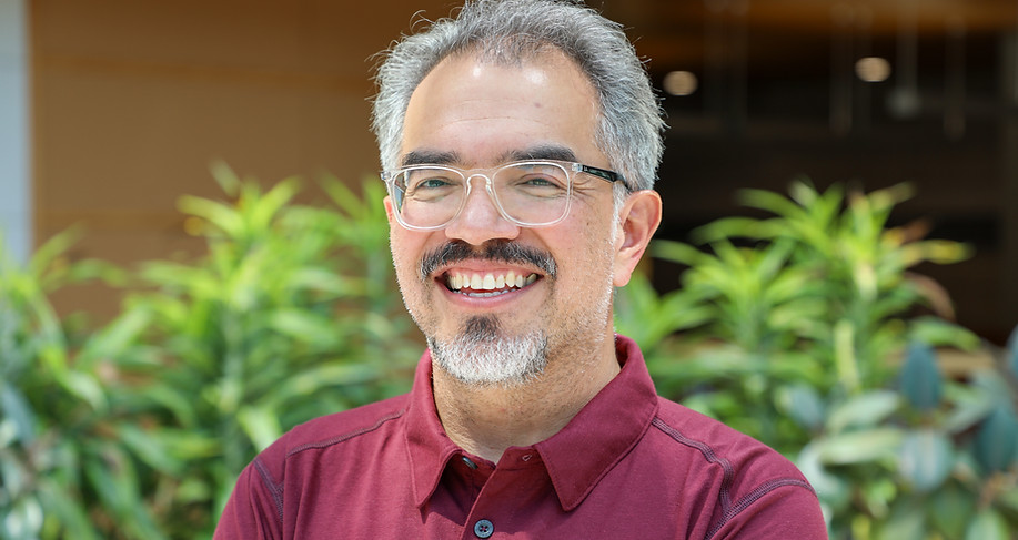 A middle-aged man with a beard and glasses smiles, standing in front of lush green plants.