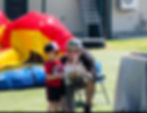 A father and son enjoy blowing bubbles together outdoors with a colorful bounce house in the background.
