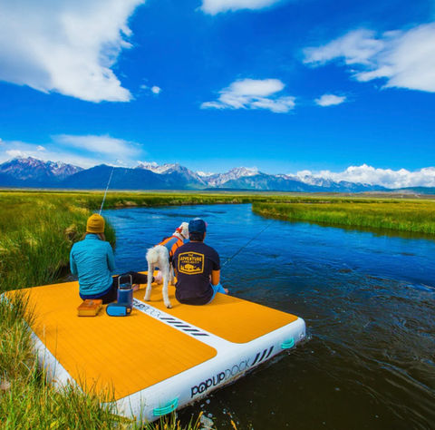 Two people sitting on an inflatable platform on a river with mountains in the background.