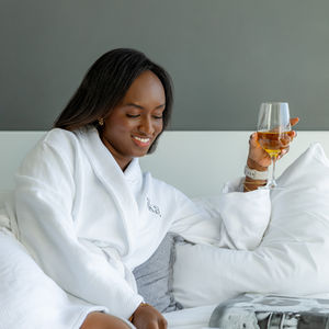 black woman in white robe sitting on the bed drinking wine and reading a magazine