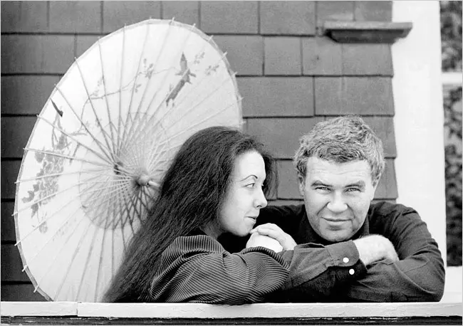 Tess Gallagher, a white woman with long dark hair is holding an umbrella and looking at her husband Raymond Carver to her left (your right). Raymond is an older white man with gray speckled hair and is wearing a dark long sleeve shirt. He is looking directly at the camera with a smirk.