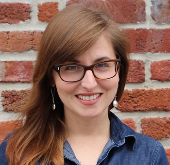 Jessica Kirkpatrick is a white woman with light brown hair. She is standing against a red brink wall and is wearing dark red glasses. She is smiling big and bright.