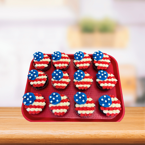Twelve cupcakes on a red tray on a wooden table, each decorated with red and white frosting stripes and blue frosting dots with a small white star, resembling the American flag. / Doce cupcakes en una bandeja roja sobre una mesa de madera, cada uno decorado con franjas de glaseado rojas y blancas y puntos de glaseado azules con una pequeña estrella blanca, asemejándose a la bandera estadounidense.