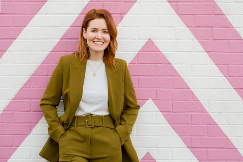 Hattie from Heck Yeah Ceremonies, stood in front of pink striped wall, wearing an olive green suit. Heck Yeah Ceremonies are an LGBTQ+ Inclusive wedding celebrant, part of Rebel Love Directory