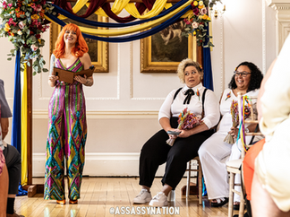 Happy couple sit down together during their wedding ceremony led by Louise from Kissass Gatherings. Louise wars a multicolored jumpsuit and has orange hair. Kickass Gatherings are an LGBTQ+ friendly supplier, part of the Rebel Love Directory.