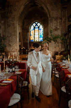 LGBTQ+ wedding photography capturing a queer couple walking hand in hand through their candlelit wedding reception at The Mount Without in Bristol. The bride wears a sparkling sequin gown, and the groom a bespoke embroidered ivory wedding suit. Surrounded by red tablescapes, gothic stained glass windows, and golden candlelight, this alternative lesbian wedding radiates romance and individuality.