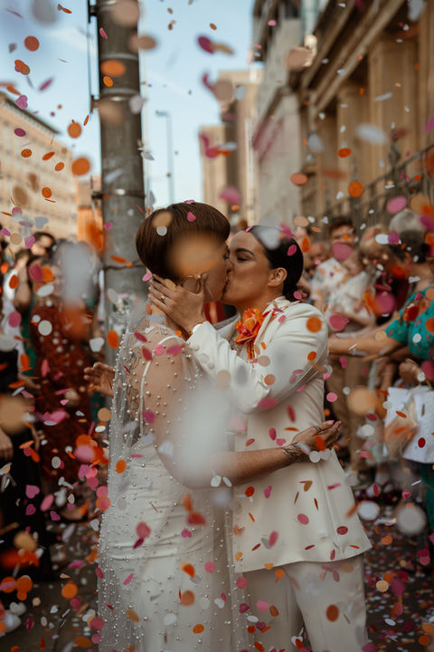 Two Brides kiss amongst a frame full of red, orange and white confetti. One is wearing a pearl covered wedding dress and the other is wearing a white wedding suit. Confetti tunnel on the streets of Liverpool just outside of Buyers Club. A bold, joyful LGBTQ+ wedding moment from an inclusive wedding photographer UK.