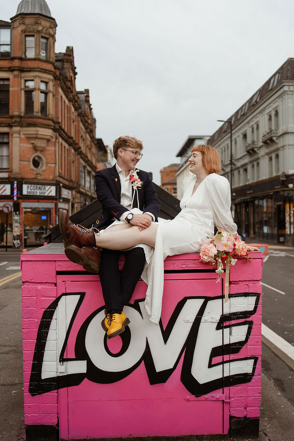 Two people sit on top of a small building in Manchester city centre which is painted bright pink with the word LOVE written across it. The people are wearing a dark wedding suit and a white wedding dress.Captured by Rebel Love Club