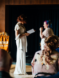 LGBTQ+ wedding ceremony led by Kickass Gatherings. The couple are both wearing white dresses and face each other during the vowel readings. Kickass Gatherings are an LGBTQ+ friendly wedding supplier, part of the Rebel Love Directory.
