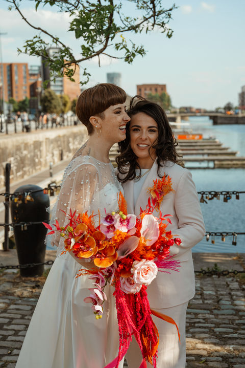 LGBTQ+ couple portrait at Liverpool’s Royal Albert Docks with bold flowers - authentic queer wedding photography UK.
