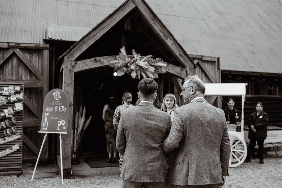 Elle and their dad wearing matching tweed suits link arms just before they walk down the aisle at The Red Barn in Norfolk, which they are stood out the front of, the photo is in black and white