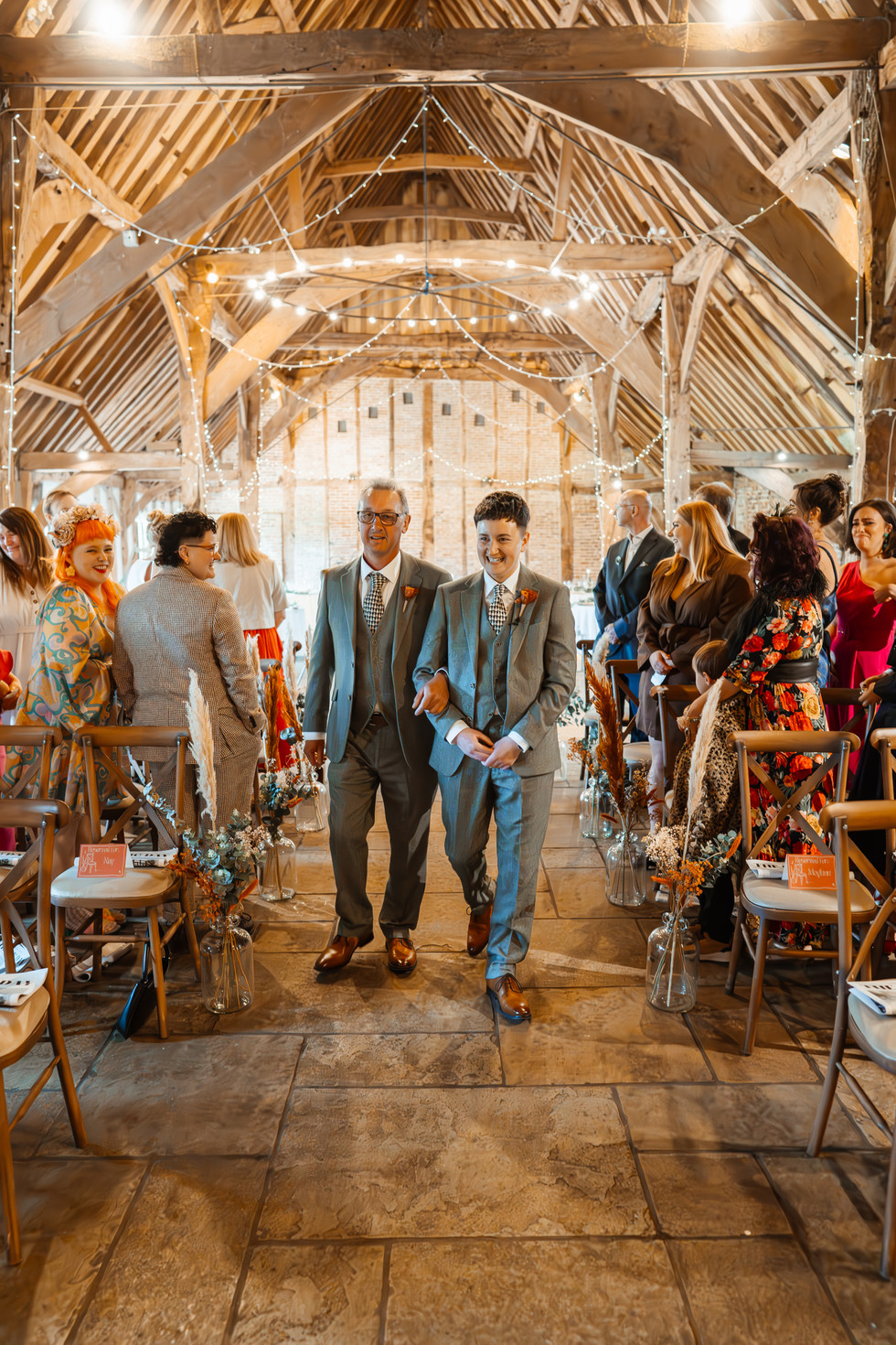 Elle and their dad walk down the aisle at The Red Barn surrounded by their best friends and family. They are wearing matching grey tweed suits and are linking arms. The feeling of emotional joy spreads across their faces
