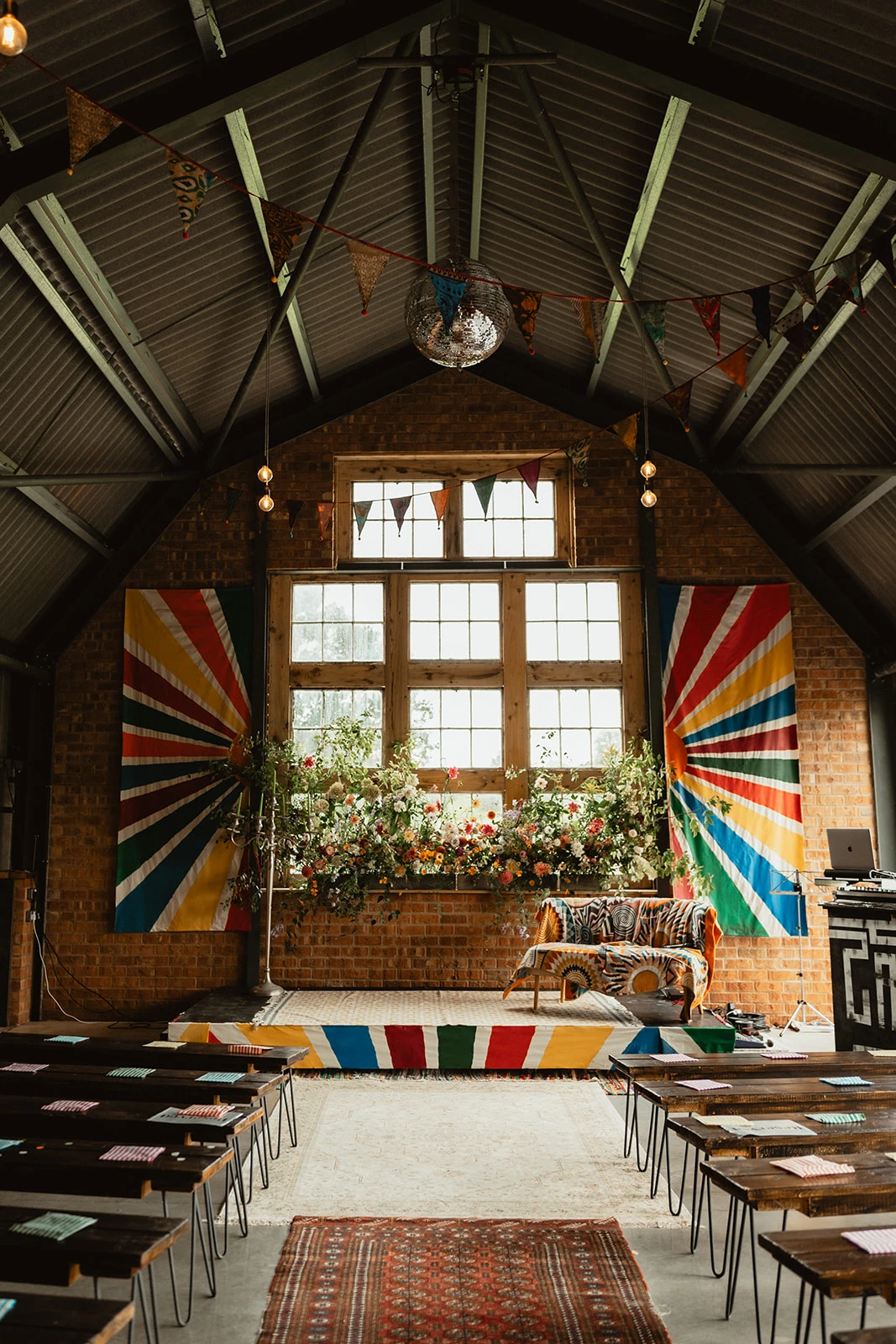 A beautifully eclectic and inclusive wedding venue, The Giraffe Shed is filled with personality. This image captures the ceremony space featuring a rustic-industrial barn setting with exposed brick walls, high metal-beamed ceilings, and large wooden windows letting in natural light. Vibrant rainbow banners frame the backdrop, with colorful floral arrangements lining the stage. Handcrafted wooden benches with patterned cushions offer seating, while warm festoon lighting and bunting create a welcoming atmosphere.