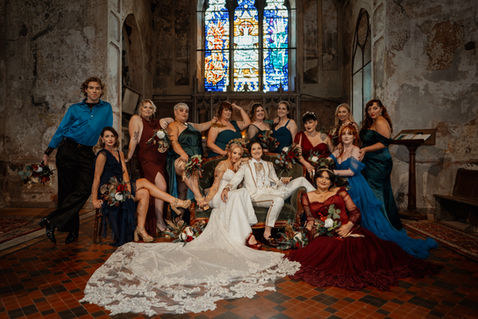 Queer wedding photography featuring a Met Gala inspired LGBTQ+ wedding party inside The Mount Without, a gothic wedding venue in Bristol. The groom in a tailored white wedding suit, the bride in a shimmering gown with a celestial crown - are surrounded by their diverse wedding party in jewel-toned outfits. A bold and inclusive feminist wedding celebration with alternative style and queer joy.