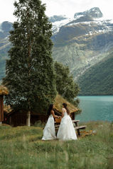 Two brides unpack their picnic surrounded by pine trees, a glacial lake and a snow capped mountain The two brides are wearing long white wedding dresses for their Norway Elopement