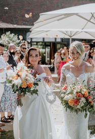 Queer, same sex couple both wearing white wedding dresses, holding hands and each holding a white and peach bouquet. Florals by Freckles and Flowers, an LGBTQ+ friendly florist, part of the Rebel Love Directory.