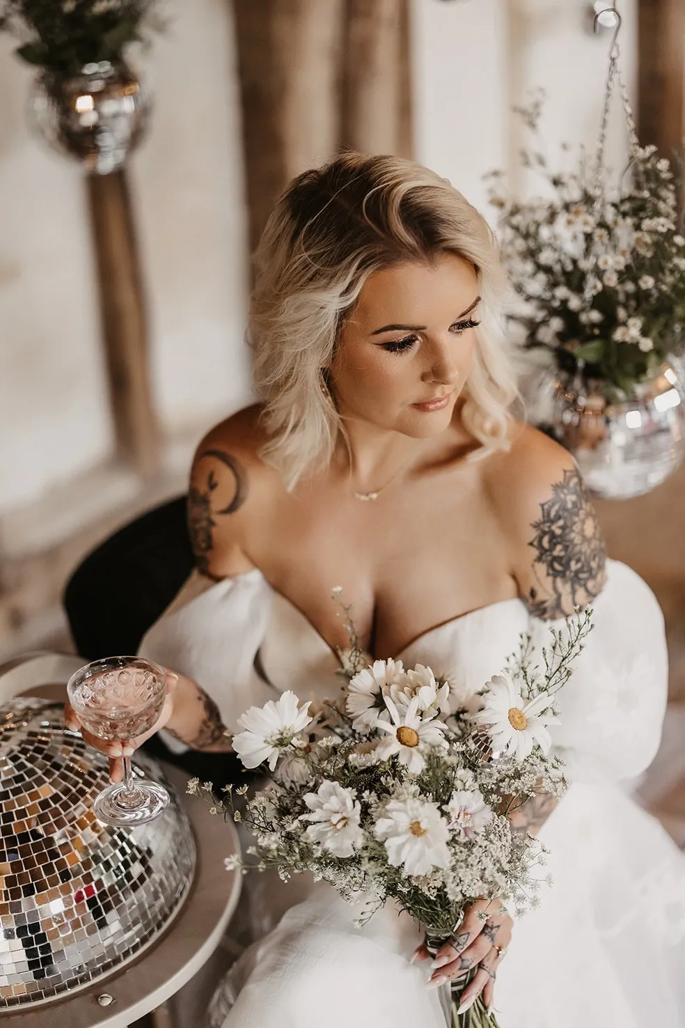 A blonde haired person in a white wedding dress, holding a glass of champagne and a bouquet. Decorations supplied by Disco Dining, an LGBTQ+ wedding supplier, part of the Rebel Love Directory.